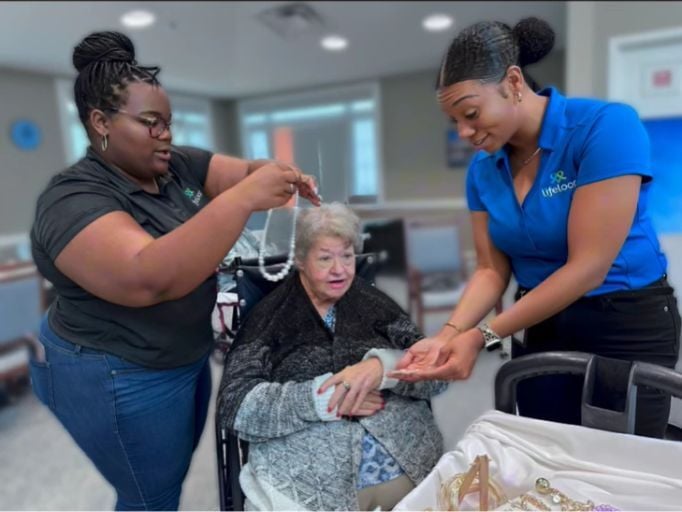 Two people helping an elderly woman in a wheelchair get ready