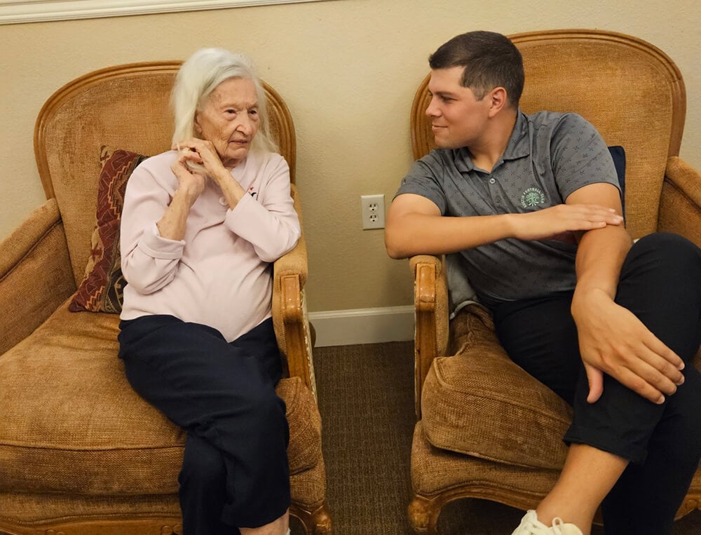 A older woman and a younger man sit in chairs and engage in conversation